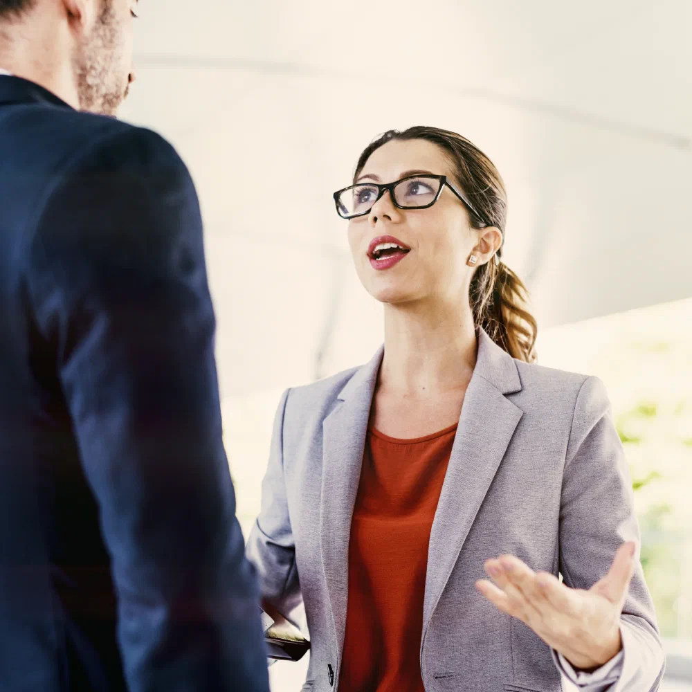 A woman in glasses and a grey blazer speaking with a colleague in a bright, modern space
